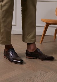 Zapatos de vestir de cuero marrón con un acabado liso y detalles texturizados, con cordones azules y un diseño de punta afilada.