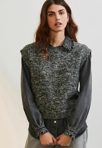 Strikket sweater-vest i grå nuancer med struktureret overflade, båret over en langærmet denimskjorte. Har en bølget krave og puffede ærmer.