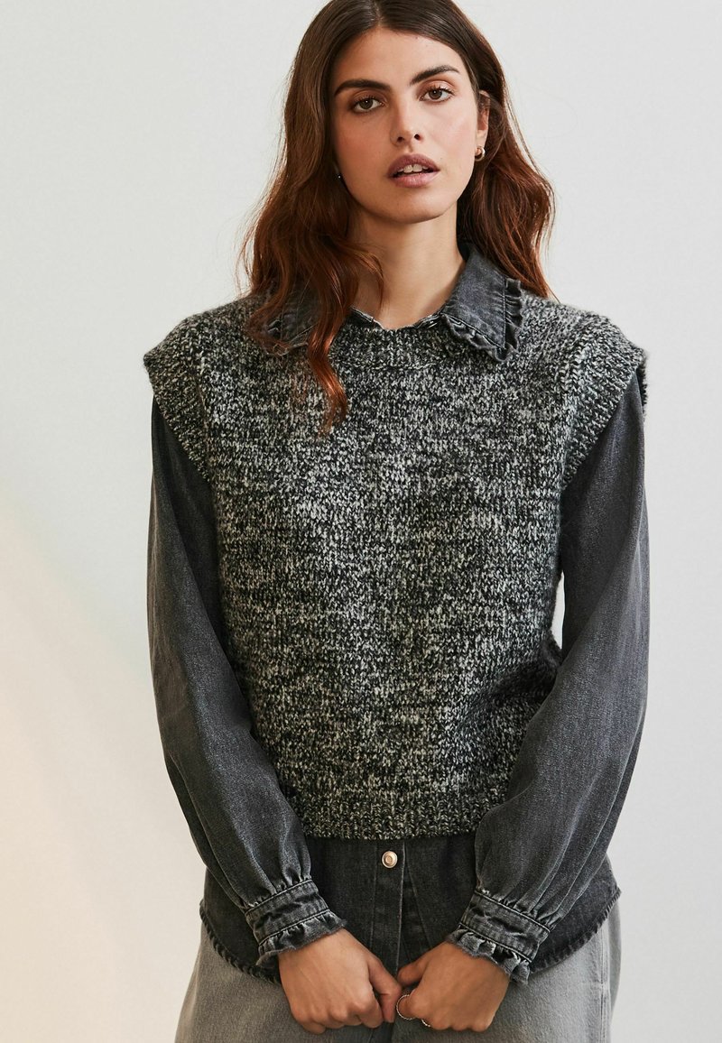 Strikket sweater-vest i grå nuancer med struktureret overflade, båret over en langærmet denimskjorte. Har en bølget krave og puffede ærmer.