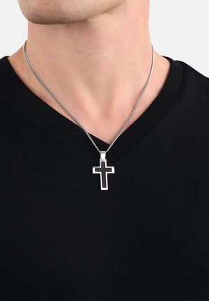 Srebrny naszyjnik łańcuszkowy z metalowym zawieszką w kształcie krzyża. Krzyż ma czarne centrum i polerowaną srebrną ramkę, noszony na czarnej koszulce z dekoltem w szpic.
