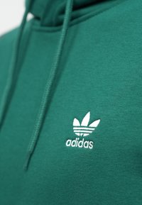 Zelená mikina s kapucňou vyrobená z mäkkej látky, s bielym vyšívaným logom Adidas a tromi pruhmi nad ním. Sú viditeľné šnúrky na stiahnutie.