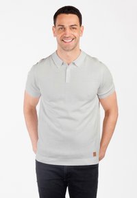 Jasnoszara koszulka polo, krótki rękaw, prążkowany kołnierzyk i mankiety, z małą brązową metką marki na dolnym szwie, noszona z ciemnymi dżinsami.
