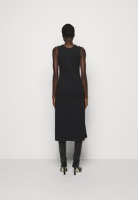 Robe midi noire sans manches cintrée portée par une personne debout de dos, associée à des bottes noires à talons montant jusqu'aux genoux.