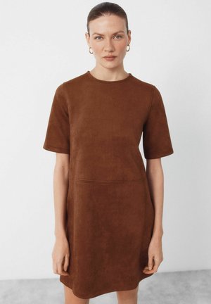 Vrouw met een lichte huid en haar naar achteren gebonden, met gouden oorringen en een bruine suède kortemouwenjurk, staand tegen een effen achtergrond.