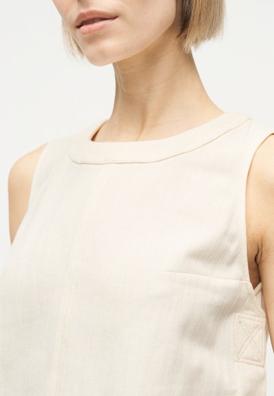 Gros plan sur une personne portant un haut sans manches de couleur beige clair avec un col rond et des détails de couture visibles près de l'épaule et de la couture latérale.