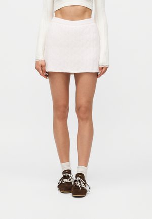 Kobieta w białej mini spódnicy z warkoczowym splotem, białym crop topie z długim rękawem, białych skarpetkach oraz brązowych zamszowych butach na sznurowadła.