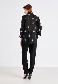 Blouse noire en voile avec broderies florales blanches, manches longues et coupe décontractée. Associée à un pantalon noir tailleur et des chaussures à talons.