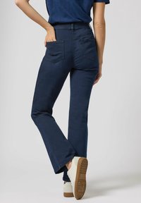 Dunkelblaue Schlagjeans mit hoher Taille, ausgestattet mit zwei Gesäßtaschen und einer glatten Stoffstruktur. Getragen mit weißen Sneakers.