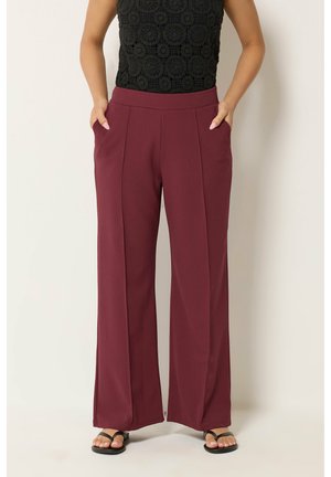 Mujer con pantalones burdeos de cintura alta y pierna ancha, top negro sin mangas de ganchillo y sandalias negras, manos en los bolsillos.