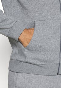 Grå hettegenser med strukturert stoff, med en sidelomme og ribbestrikkede mansjetter. Designet inkluderer glidelåslekking foran.