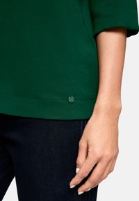 Grünes Baumwollhemd mit abgerundetem Saum und sichtbaren Nähten; verfügt über ein kleines grünes Knopfdetail in der Nähe des Saums, kombiniert mit Denim-Stoff.