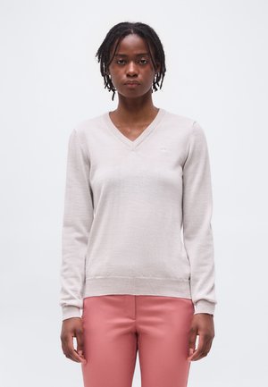 Γυναίκα με κοντά πλεγμένα μαλλιά φοράει ένα ανοιχτό μπεζ πουλόβερ με V λαιμόκοψη και ψηλόμεσα παντελόνια σε σκούρο ροζ χρώμα, στέκεται μπροστά σε μια μονόχρωμη επιφάνεια.