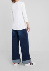 Person som bär vida, mörkblå, uppvikta jeans, en vit topp med trekvartsärmar och vita sneakers med flerfärgade ränder, står på sidan.