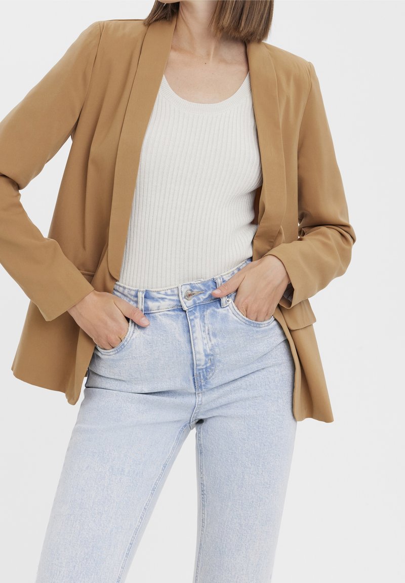 Blazer marron à texture lisse, top sans manches à côtes légères, et jean bleu clair taille haute. Le mannequin pose les mains dans les poches.