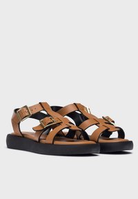 Sandalias de cuero marrón con diseño entrelazado, dos hebillas ajustables y una suela de goma negra para mayor tracción. Acabado suave con los dedos de los pies al aire.