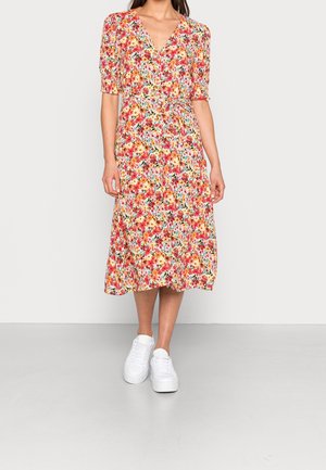 Femme portant une robe fleurie mi-longue avec des manches bouffantes et un décolleté en V, associée à des baskets compensées blanches, debout sur un sol uni.