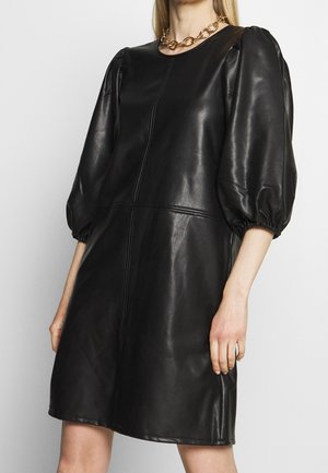 Femme portant une robe noire en similicuir avec des manches bouffantes jusqu'au coude et un collier en chaîne dorée, debout devant un fond uni.