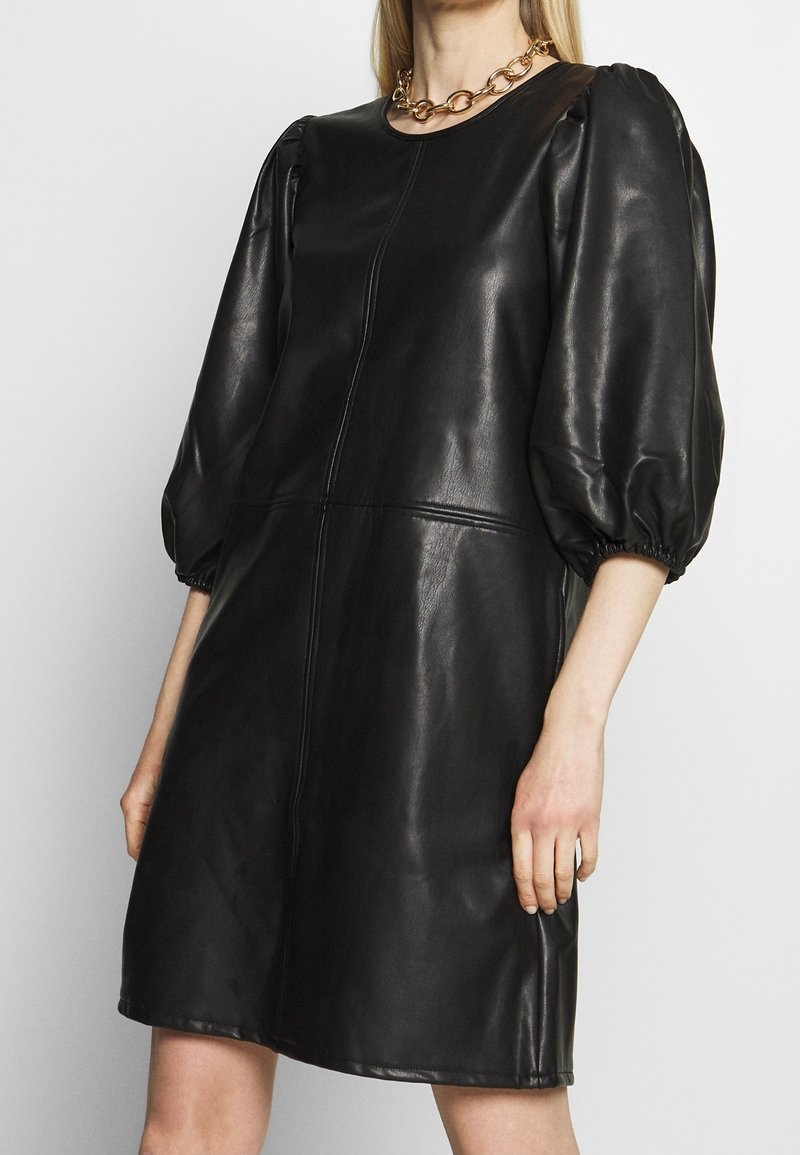 Femme portant une robe noire en similicuir avec des manches bouffantes jusqu'au coude et un collier en chaîne dorée, debout devant un fond uni.