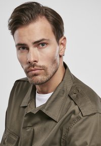 Camisa verde oliva de botones con cuello y bolsillo de parche. El material parece suave con un ligero brillo. Llevada sobre una camiseta blanca.