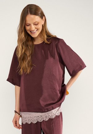 Femme aux longs cheveux portant un haut bordeaux ample et un pantalon avec un ourlet en dentelle, souriante et regardant vers le bas, mains détendues sur les hanches.