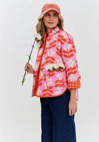 Svetlo ružový kvetinový kabát s oranžovými vzormi listov, so zipsom, bočnými vreckami a kockovaným akcentom na manžetách. Nosený s tmavými džínmi.