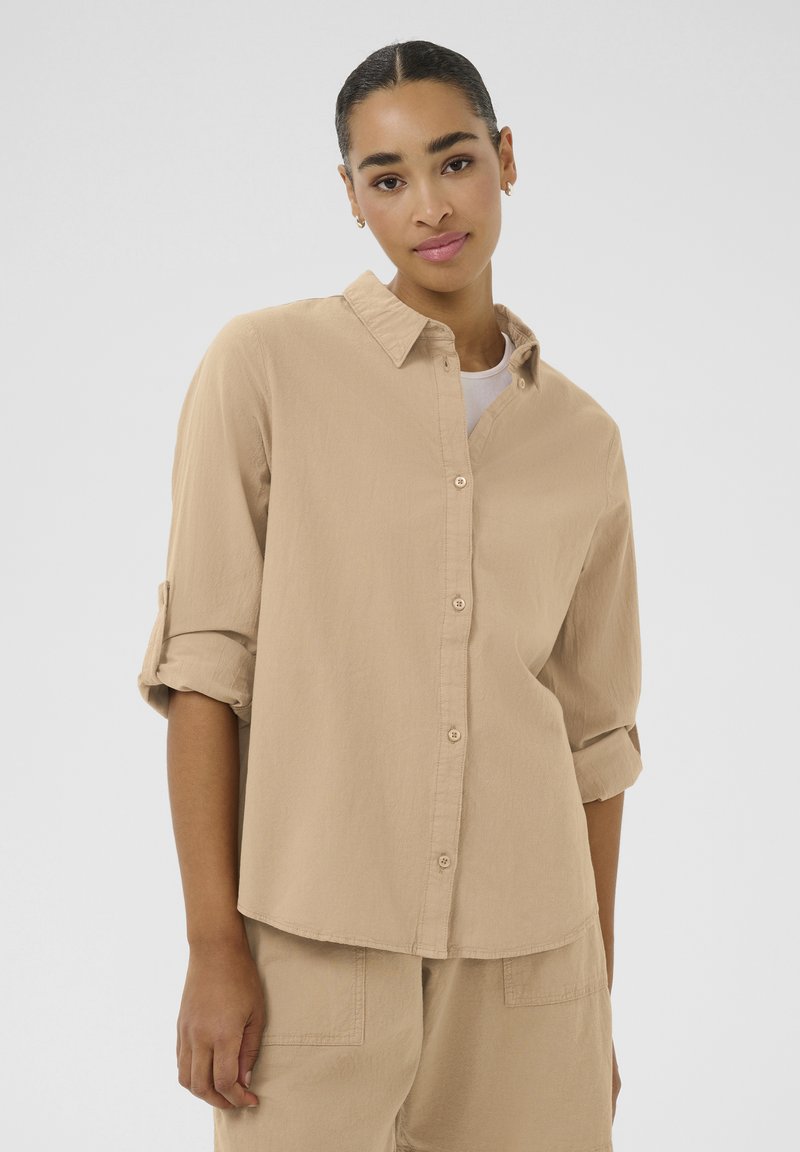 Beige knoopshirt met een kraag, lange mouwen opgerold tot de ellebogen, een ontspannen pasvorm en een zachte textuur. Draag over een wit shirt.