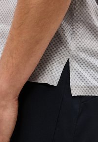 Ανοιχτό γκρι κοντομάνικο πουκάμισο με υφή και πλευρική σχισμή στο τελείωμα πάνω από μαύρο παντελόνι, με κοντινό πλάνο στο αντιβράχιο και το χέρι ενός ατόμου.