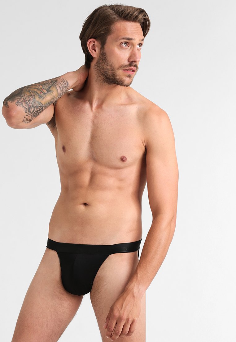 Perizoma nero indossato da un modello maschile, realizzato in un tessuto liscio, con cintura elastica e copertura minimale, esposto su uno sfondo neutro.