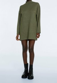 Henkilö, joka on pukeutunut oliivinvihreään pitkähiharaiseen mini-alaan, jota on paritettu mustilla paksuilla nilkkabootseilla tasaisessa taustassa.