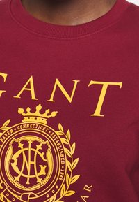 Sudadera color burdeos con texto amarillo en negrita "GANT" y un emblema circular con una corona, establecido en 1949, rodeado de hojas de laurel.