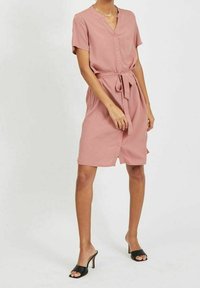Růžové košilové šaty s krátkými rukávy, opásané v pase a s náprsní kapsou. Hladký materiál, délka do poloviny stehna, doplněné černými lodičkami na podpatku.
