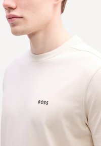 Beige, rövid ujjú póló, kerek nyakú, bal mellen fekete "BOSS" felirattal. Simító, könnyű anyag.