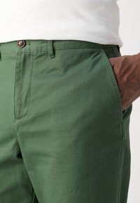 Pantaloni di cotone verdi con una texture liscia, caratterizzati da una chiusura con bottone, tasche frontali e una tasca laterale con interno marrone.