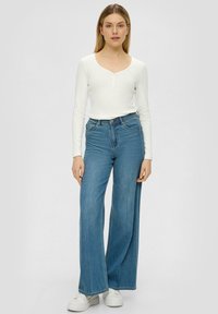 Witte langslavige top met een ronde hals en knoopdetail, gecombineerd met lichtblauwe high-waisted wijdpijpjens en witte sneakers.