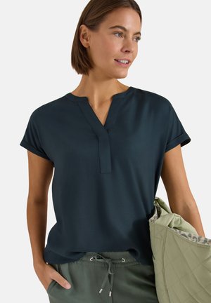 Femme portant un haut bleu marine à manches courtes et col en V, ainsi qu’un pantalon olive, tenant un sac matelassé vert clair avec une doublure imprimée animal.