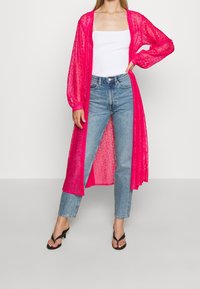 Kardigan pitkä, läpinäkyvä, kuuman vaaleanpunainen, jossa on pitsikuvio ja löysät hihat, yhdistetty valkoiseen topiin ja vaaleansinisiin korkeavyötäröisiin farkkuihin. Mustat korkokengät.