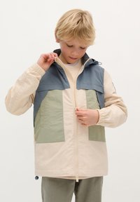Veste légère tricolore avec un corps beige, un haut bleu foncé et des patchs verts olive, dotée d'une fermeture éclair à l'avant et d'une capuche.