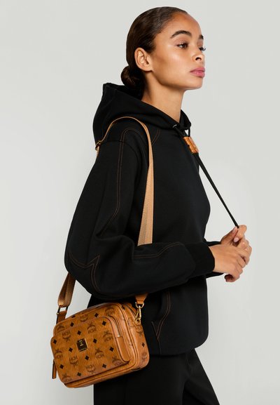 Femme en sweat à capuche noir porte un sac bandoulière à motifs marron avec une sangle réglable sur fond clair.