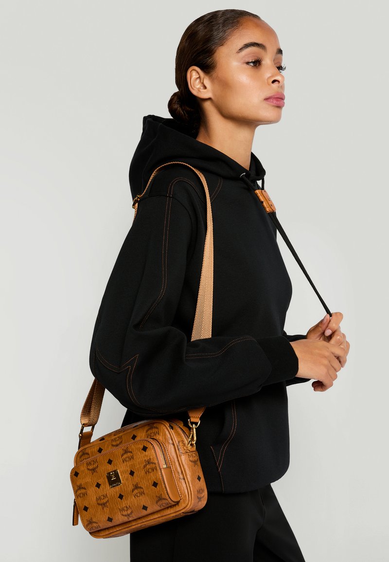 Frau in schwarzem Hoodie trägt eine braun gemusterte Umhängetasche mit verstellbarem Riemen vor einem hellen Hintergrund.