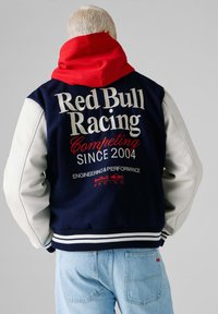 Person med kort blondt hår iført en marineblå og hvid Red Bull Racing-jakke over en rød hættetrøje, kombineret med lyseblå jeans.