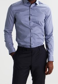 Lichtblauw overhemd met kleine, donkere geometrische patronen, lange mouwen, kraag en knoopsluiting aan de voorkant, gecombineerd met donkere broek.