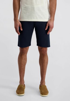 Mann trägt marineblaue knielange Shorts, ein hellbeiges T-Shirt und braune Wildleder-Slipper, steht vor einem schlichten Hintergrund.