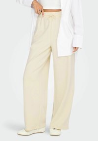 Žena v béžových širokých kalhotách s šňůrkou v pase, bílém cropped topu, oversized bílém tričku a bílých teniskách na světlém pozadí.
