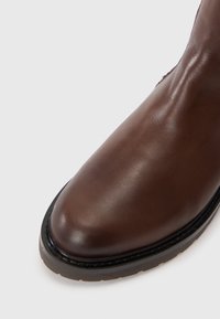 Bottine en cuir marron avec une finition lisse, bout arrondi, semelle noire robuste et détails de couture discrets le long du bord.
