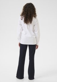 Camicia bianca a maniche lunghe, taglio dritto, vista posteriore; abbinata a jeans in denim scuri a zampa d'elefante. Tessuto liscio, design semplice, senza motivi visibili.