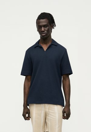 Άνδρας με ντρέντλοκς, που φοράει μπλε κοντομάνικο πόλο και μπεζ σορτς με λευκές κάθετες ρίγες, στέκεται μπροστά σε απλό φόντο.