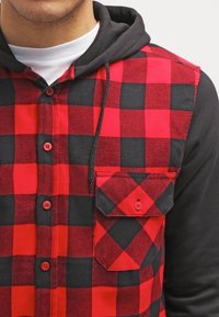 Camicia di flanella a quadri rossi e neri con cappuccio, caratterizzata da un colletto con bottoni, tasca frontale e maniche nere a contrasto.