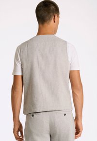 Hellgraue, ärmellose Weste aus strukturiertem Stoff. Sie hat einen geraden Schnitt mit einer Rückennaht und wird über einem weißen T-Shirt getragen.