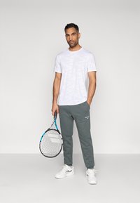 Homem vestido com uma t-shirt branca, calças de moletom cinzas e ténis brancos, segurando uma raquete de ténis, de pé contra um fundo claro e liso.