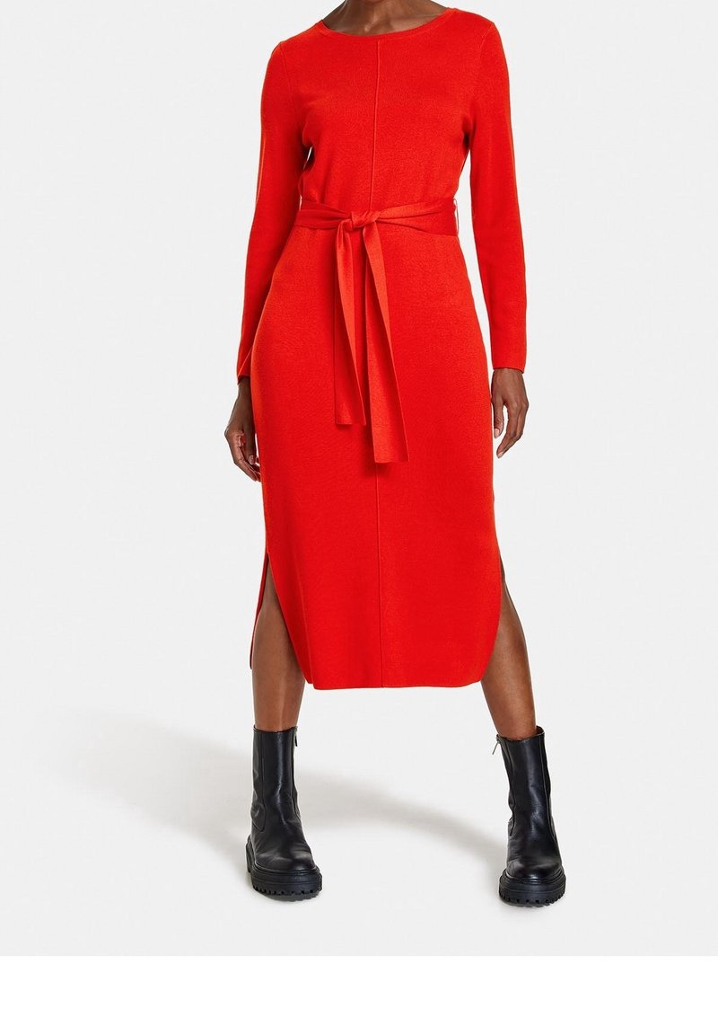Robe rouge à manches longues avec une taille cintrée, fentes latérales et une ceinture assortie. Portée avec des bottines noires à la cheville. Texture lisse.